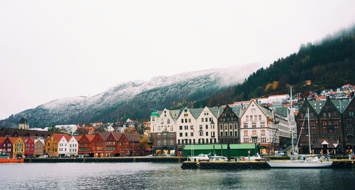 Hvite bygg på en brygge med vann i forgrunnen og fjell i bakgrunnen.