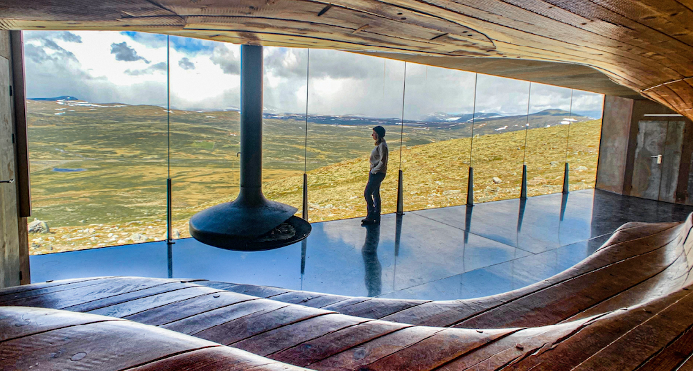 Bilde av en person som staar inne i et bygg og ser ut av store vinduer som dekker hele flaten.